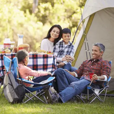 A family of four enjoys a picnic by their tent in a sunny, green park. They are smiling and relaxed, with food and drinks on a plaid tablecloth.